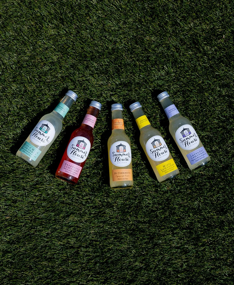 Five bottles of Summer House drink on green grass, each with different flavored labels, including lemon, raspberry, and lavender lemonade.