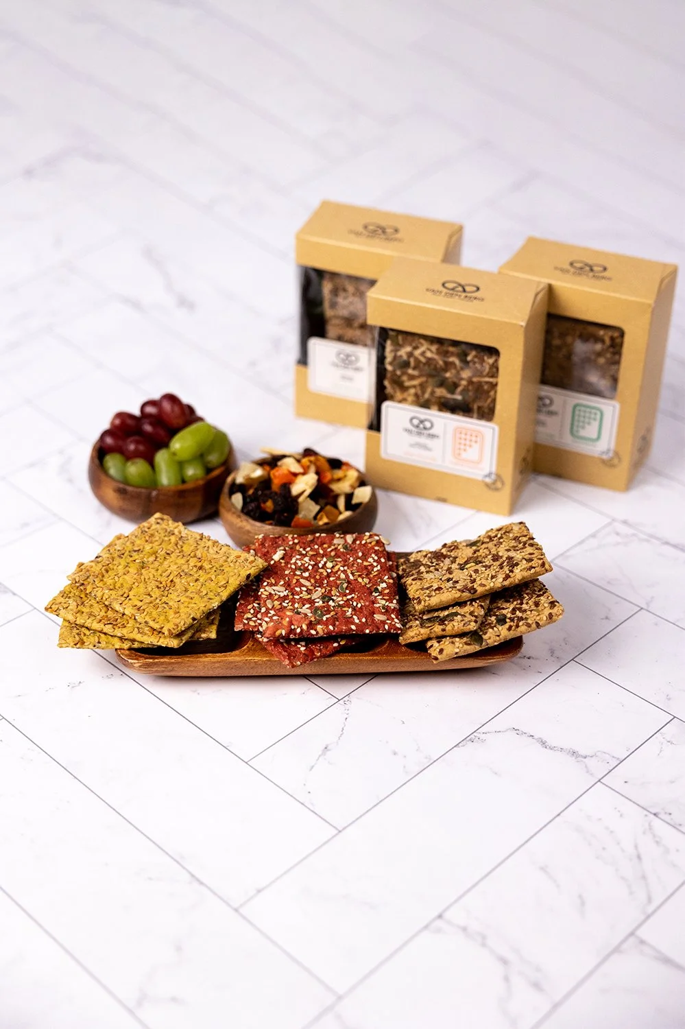 Assorted snack crackers, bowls of grapes and dried fruit, and boxes of tea on a white tiled surface.