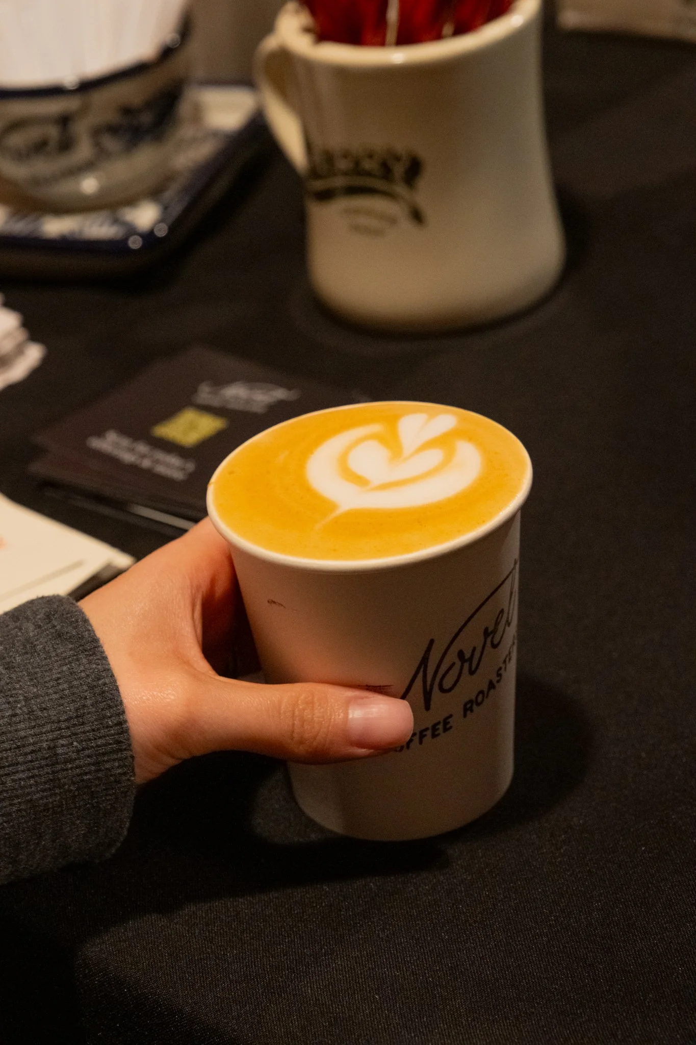 A person holding a cup of latte with heart-shaped latte art on top, on a black table with a white mug and black napkins in the background.
