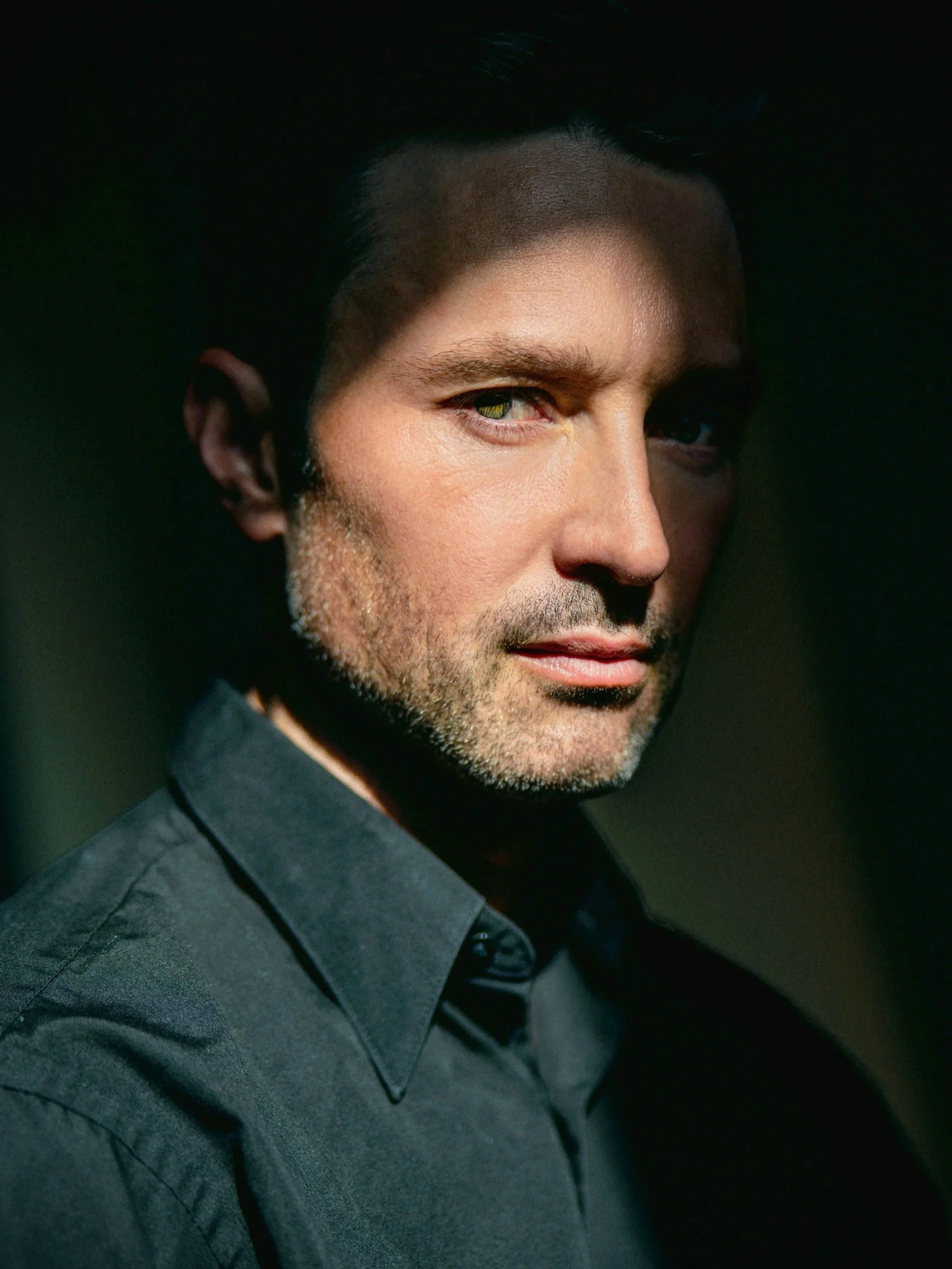 Close-up portrait of a man with dark hair, stubble, and light skin, wearing a dark shirt, looking at the camera with a serious expression, in a dimly lit environment.
