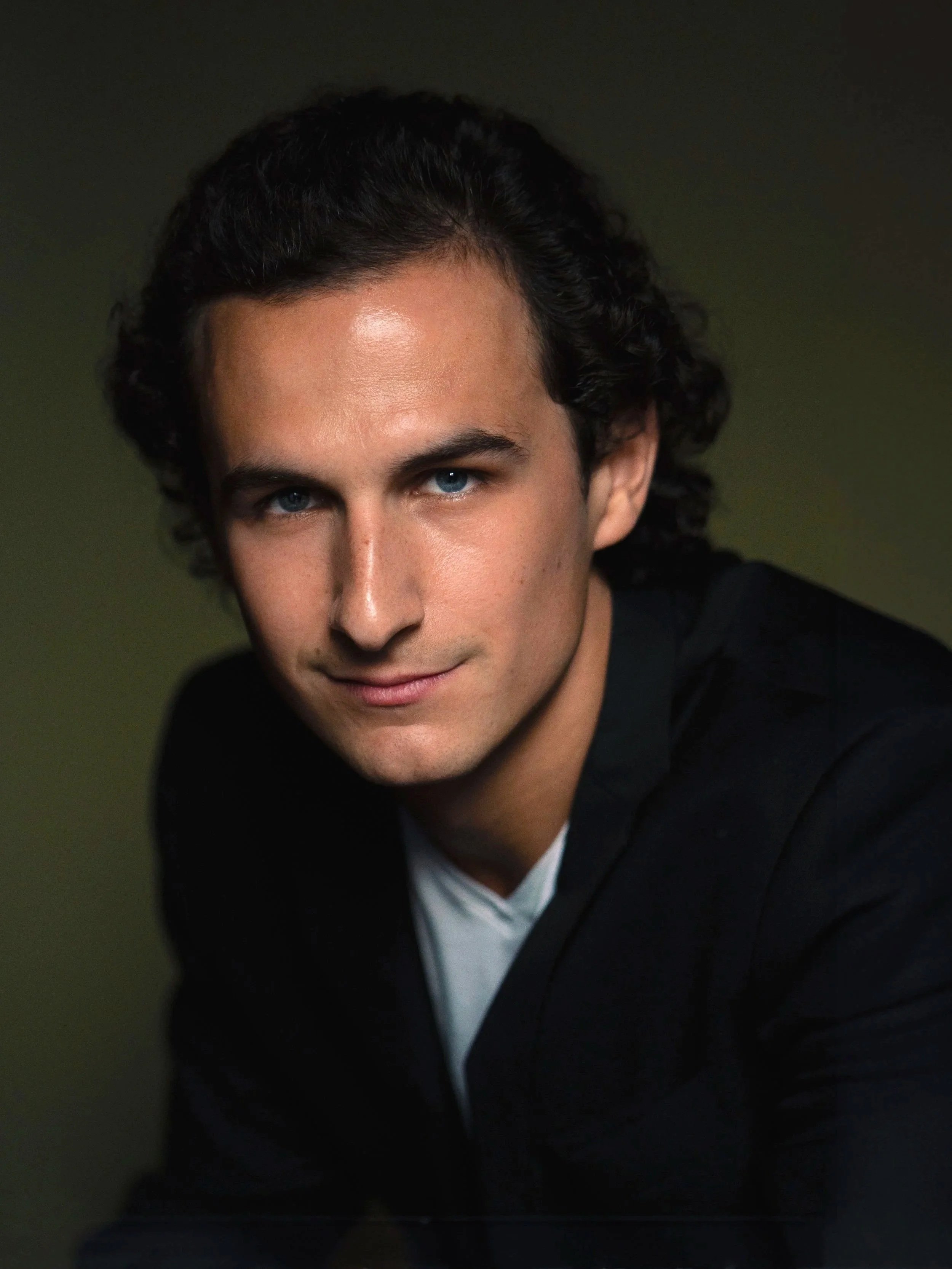 A close-up portrait of a young man with blue eyes, dark curly hair, wearing a black jacket and a white shirt, looking directly at the camera against a dark background.