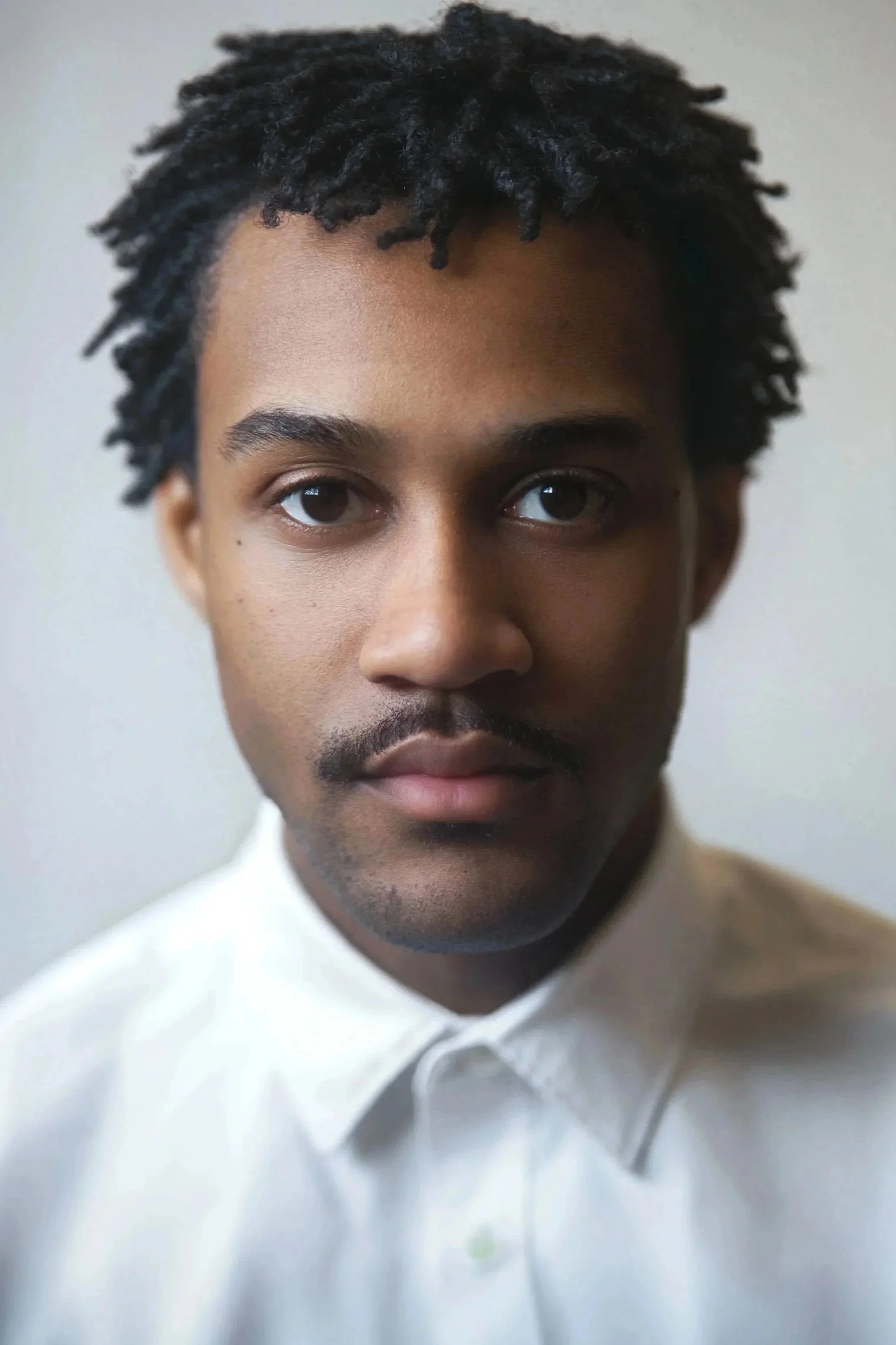 A close-up portrait of a young man with short twisted black hair, medium skin tone, wearing a white collared shirt, looking directly at the camera with a neutral expression.