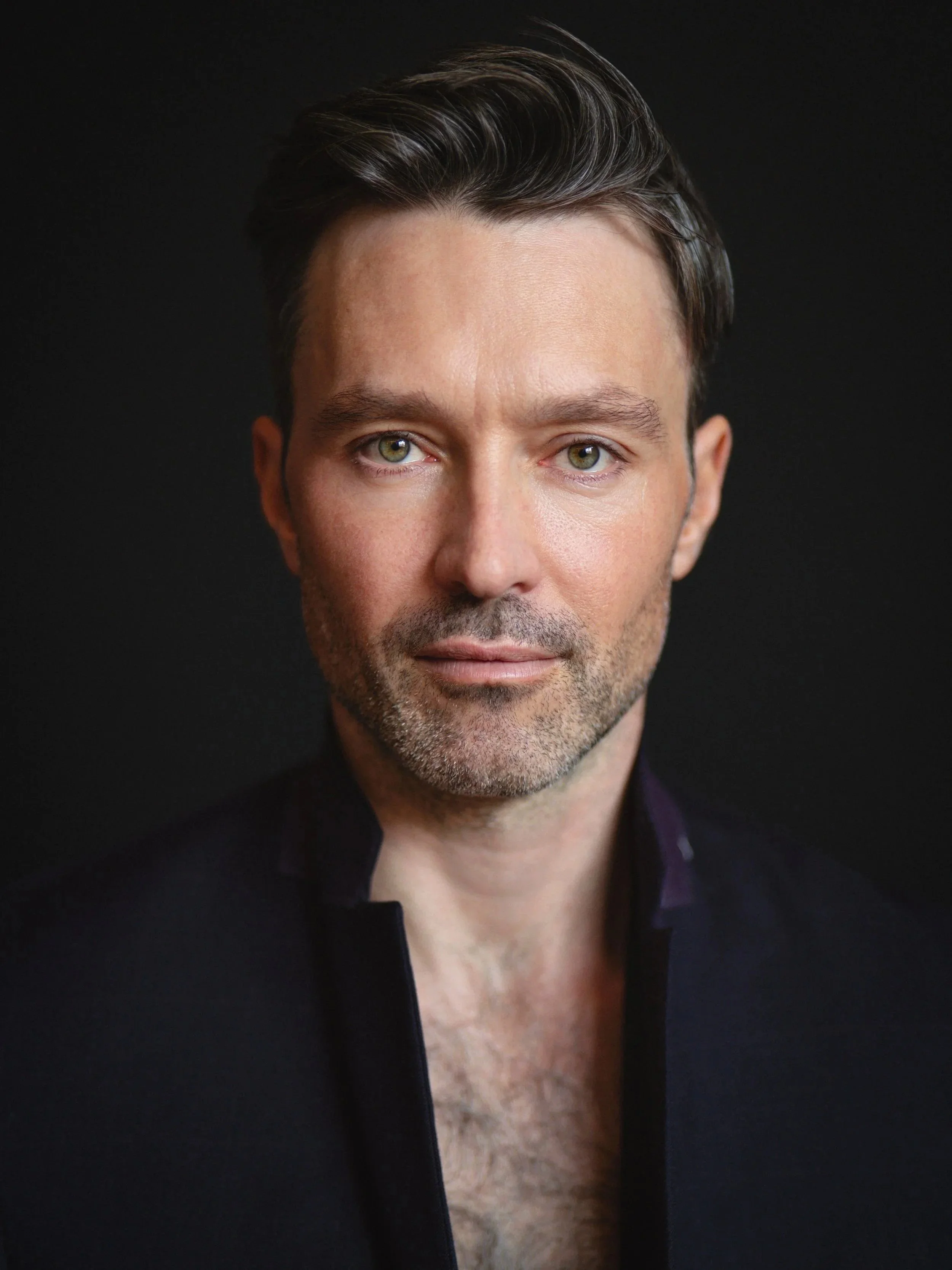 Close-up portrait of a man with short dark hair, light skin, and green eyes, wearing a dark jacket against a black background.