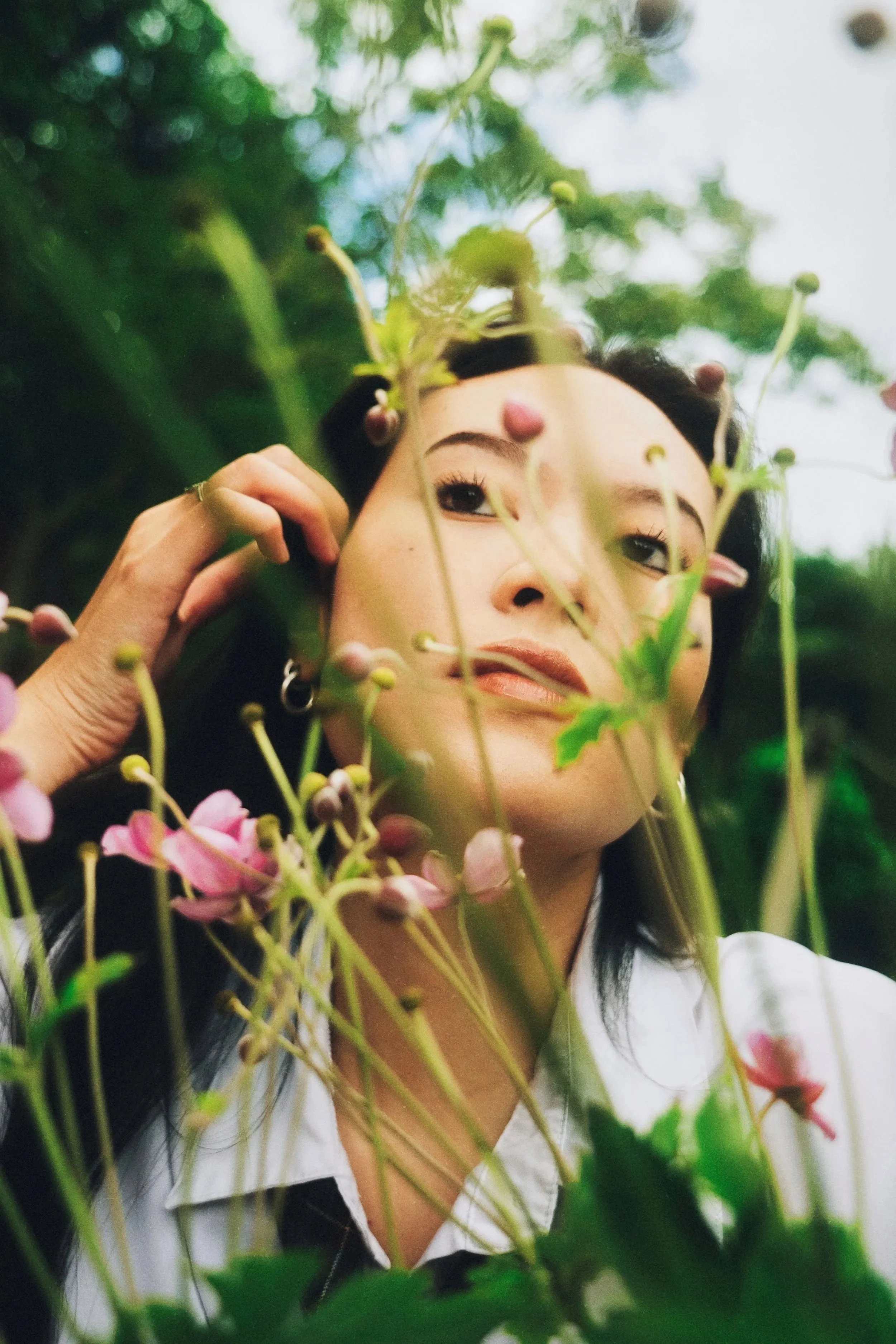 A woman with dark hair and earrings poses among flowering plants, looking slightly upward with a soft expression.
