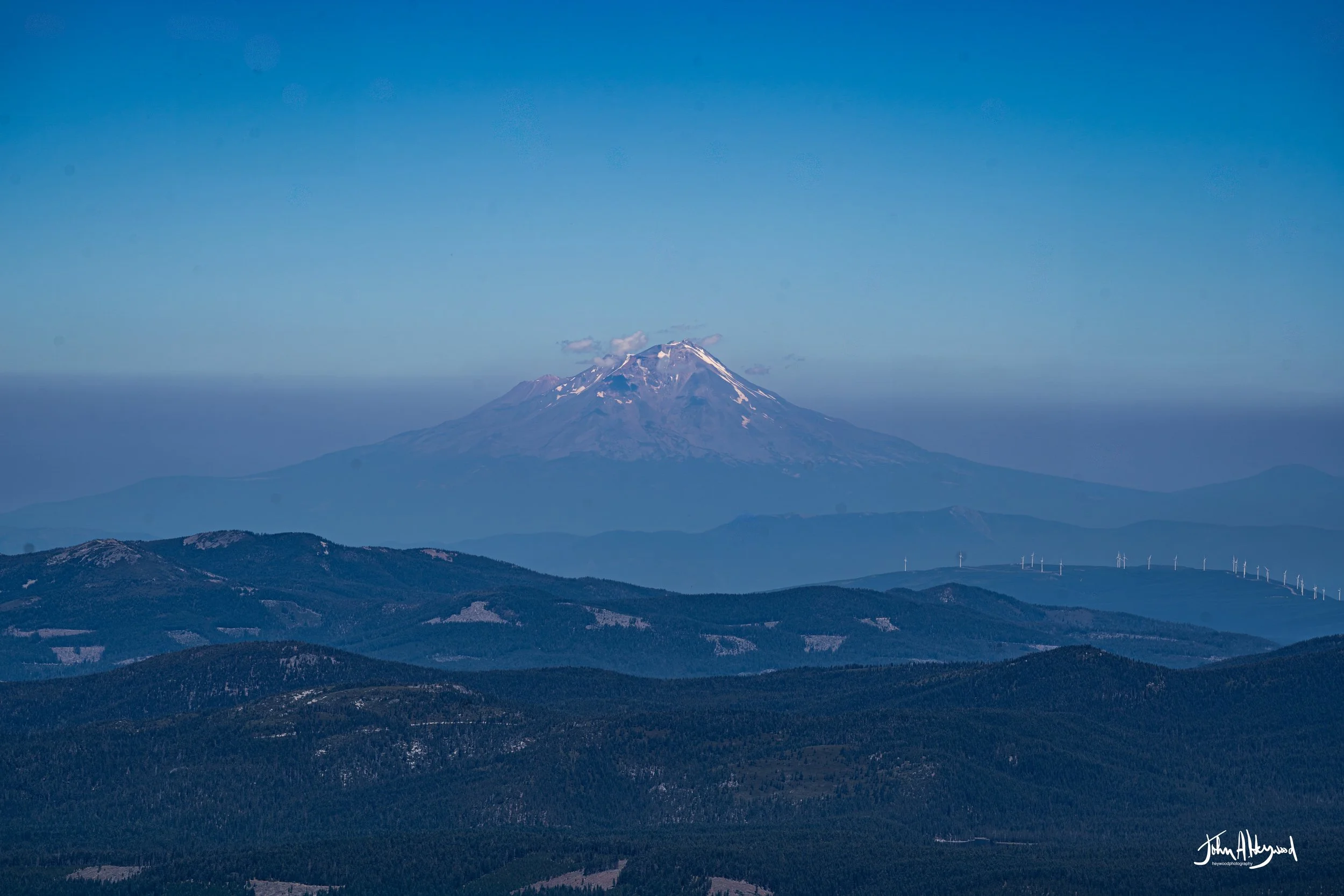 A mountain with snow on its peak, surrounded by hills and wind turbines, under a clear blue sky.