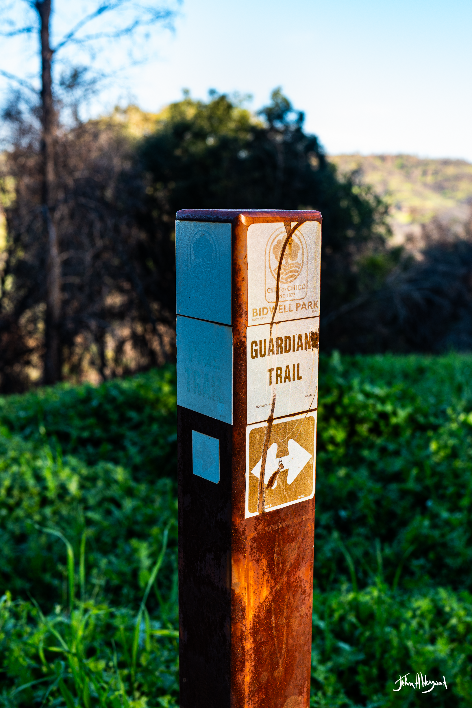 A rusted trail marker post for Bidwell Park's Guardian Trail, with an arrow pointing to the right, in a green outdoor setting with trees and hills in the background.