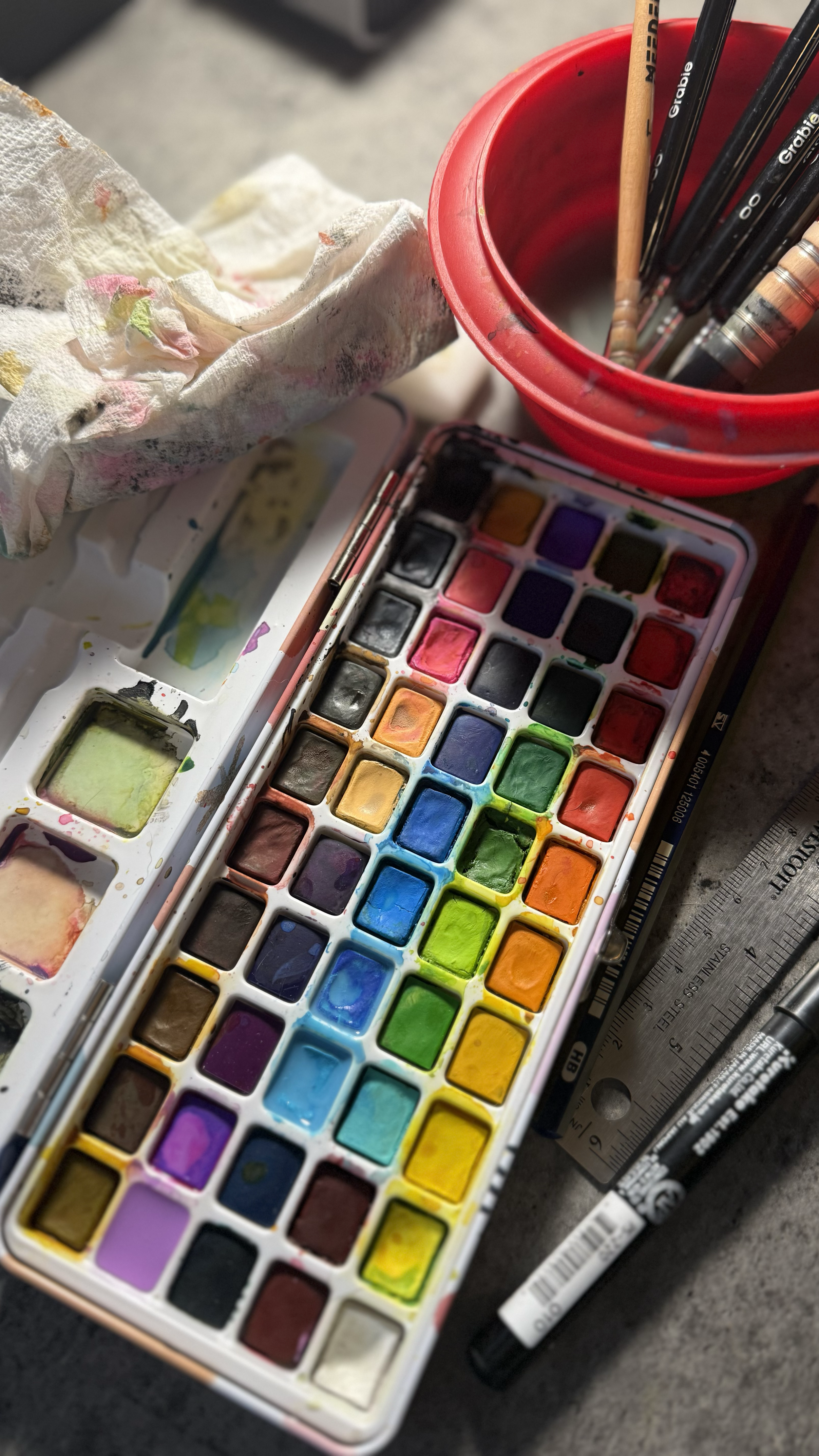 Watercolor paint set with multiple colors, paintbrushes, a pencil, and a ruler on a gray surface.