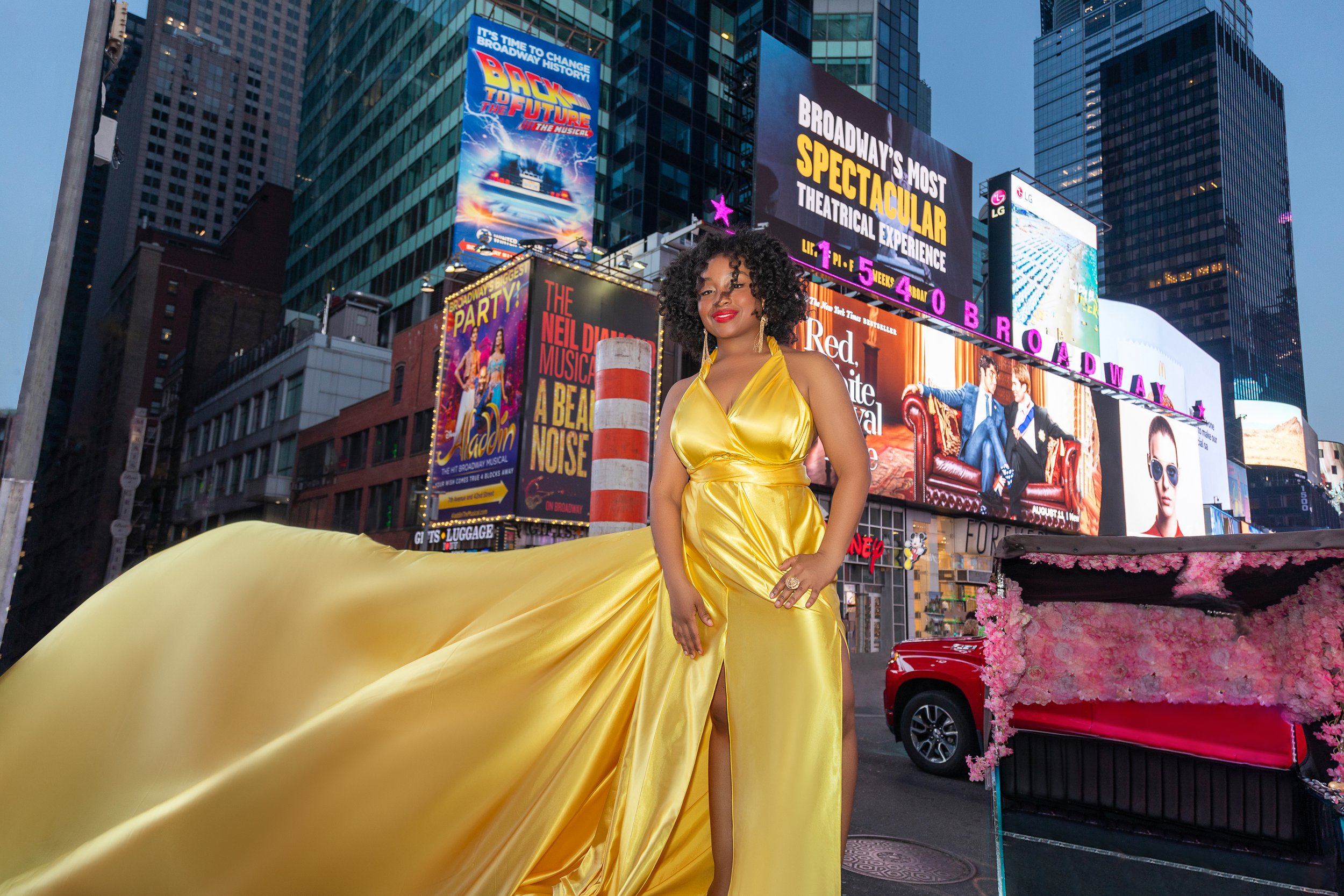 A person in a flowing yellow gown standing in Times Square, New York City, surrounded by colorful Broadway billboards at dusk.