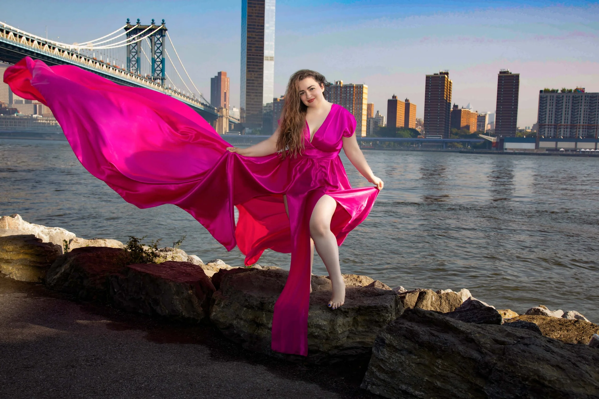Person in a flowing pink flying dress standing on riverbank with a bridge and Manhattan city skyline in the background.