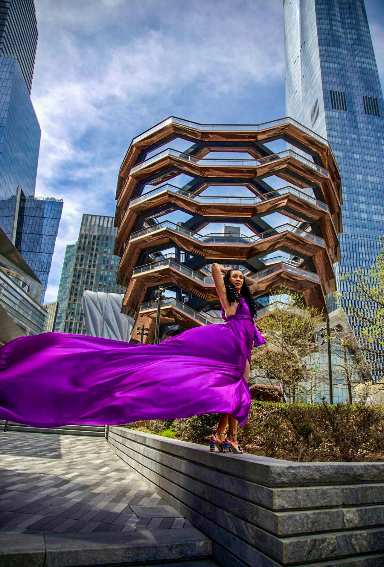Woman in a flowing purple dress stands in front of the Vessel structure in New York City, with skyscrapers in the background.