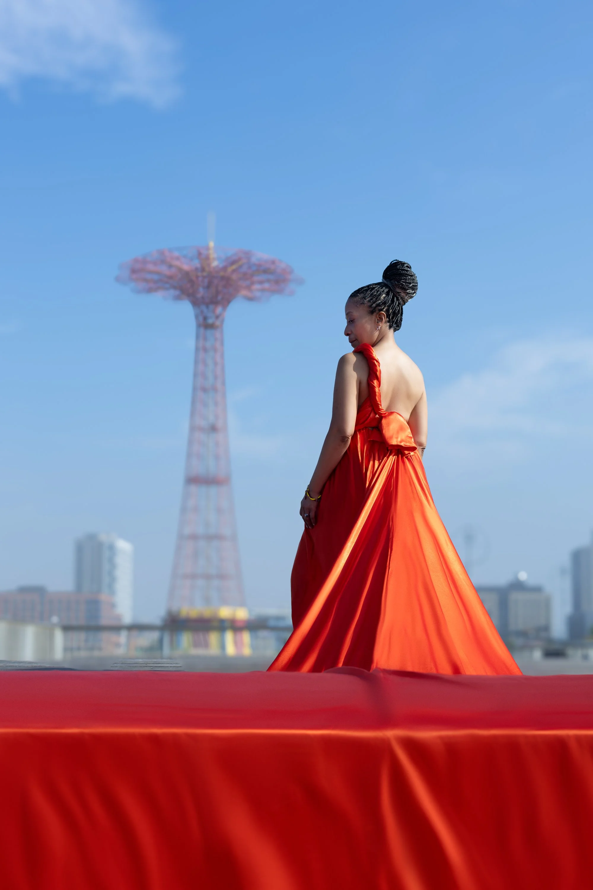 Woman in red dress standing near Coney Island Parachute Jump tower with city skyline in background.