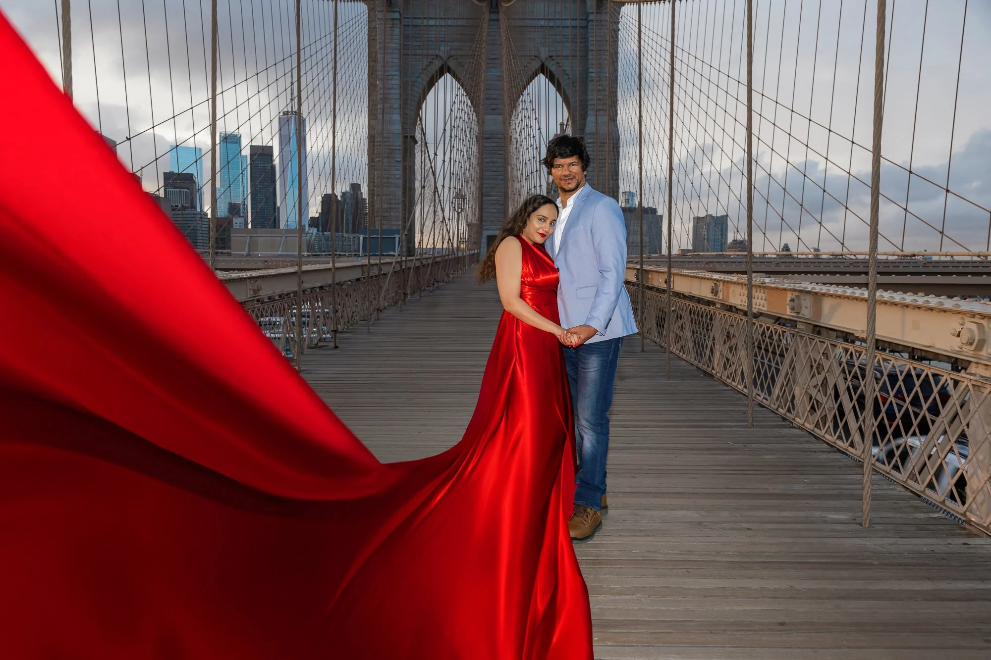 A couple posing on the Brooklyn Bridge, with the woman in a flying red dress and the man in a blue jacket.