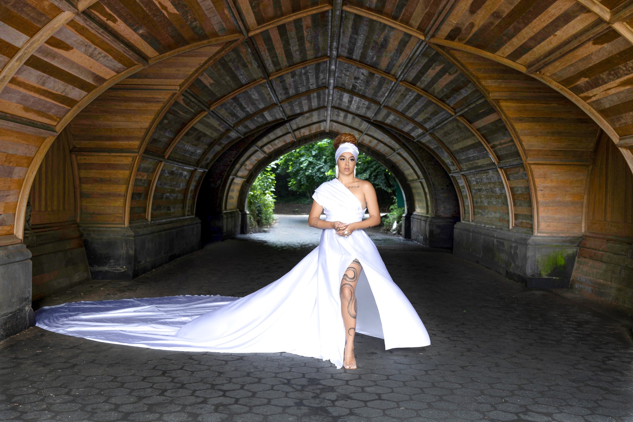 Woman in a long white gown with a slit, standing in a wooden arched tunnel, with tattoos visible on her leg.