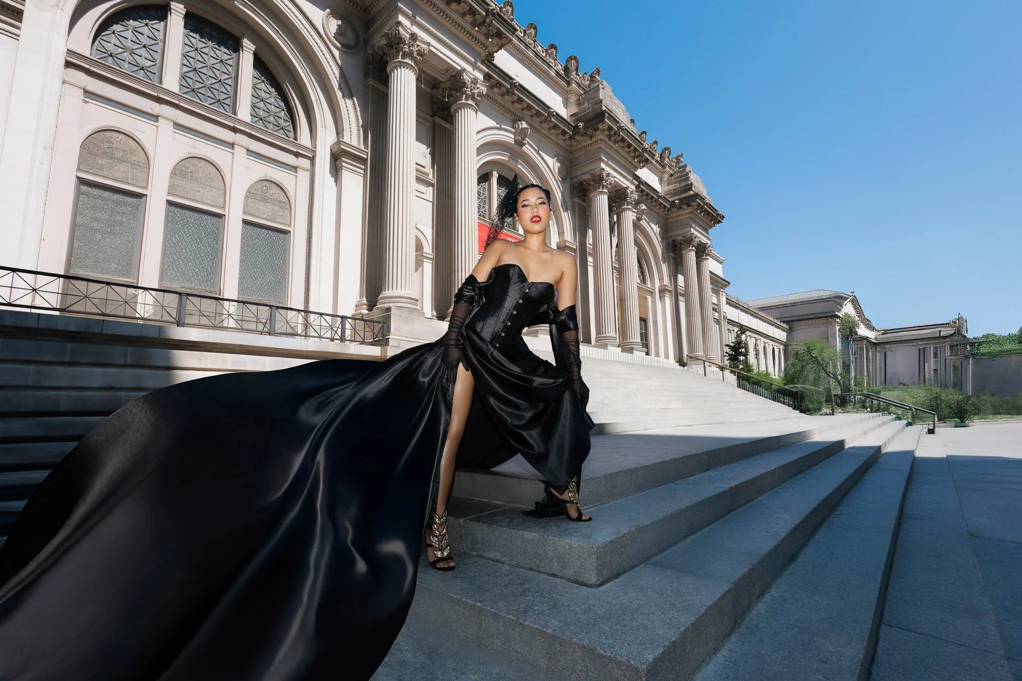 A woman in a black flying dress with a high slit, black gloves, and high heels standing on steps of the Met on a sunny day.