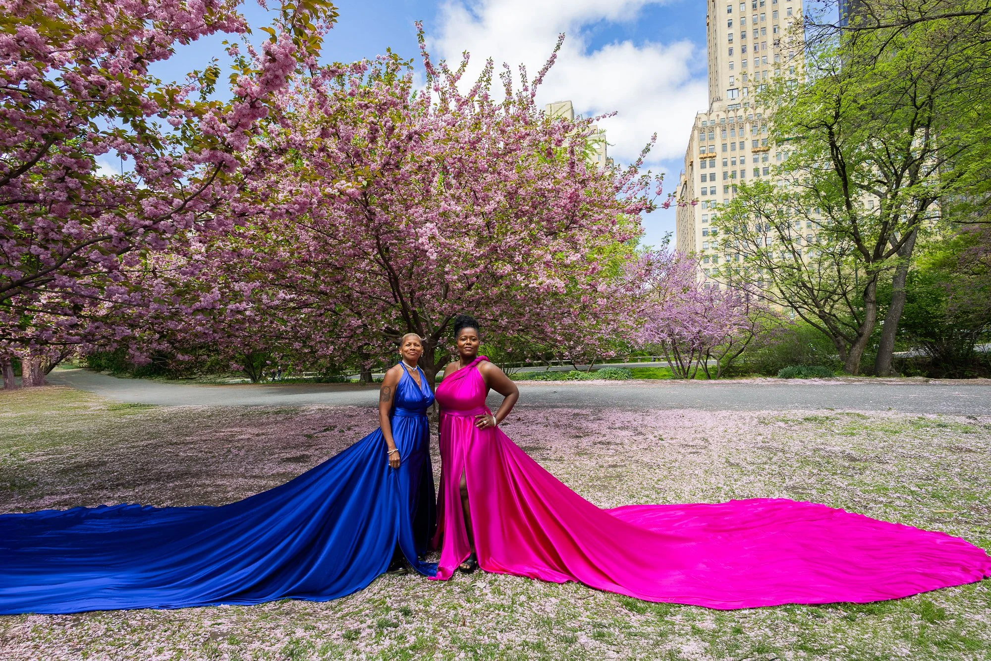 Two women in long, flowing, brightly colored dresses standing in front of blooming pink cherry blossom trees in a city park, with tall buildings in the background.