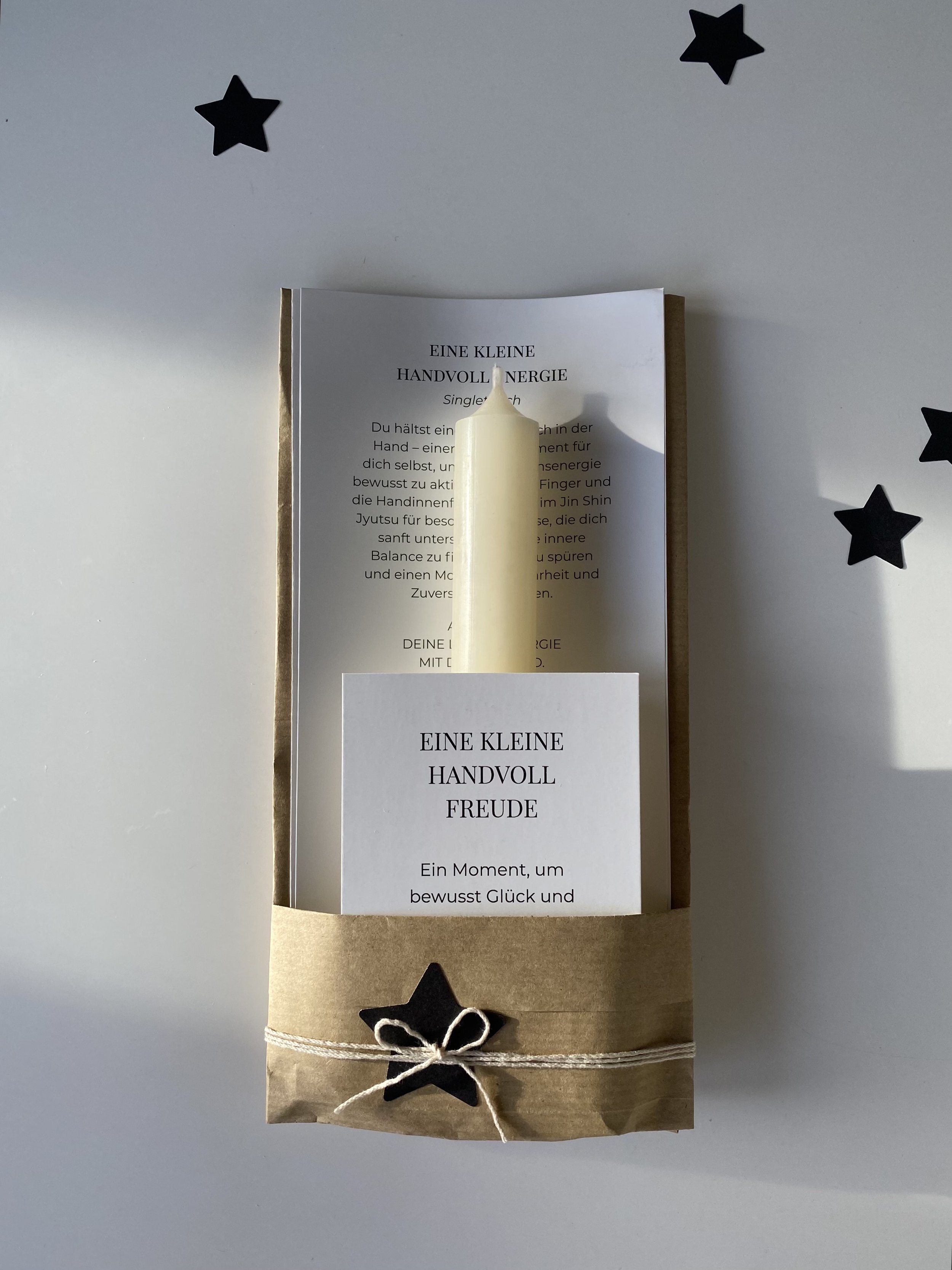 Ein braunes Papierpaket mit weißem Band und schwarzen Sternen, darin eine Kerze, auf einem weißen Tisch.
