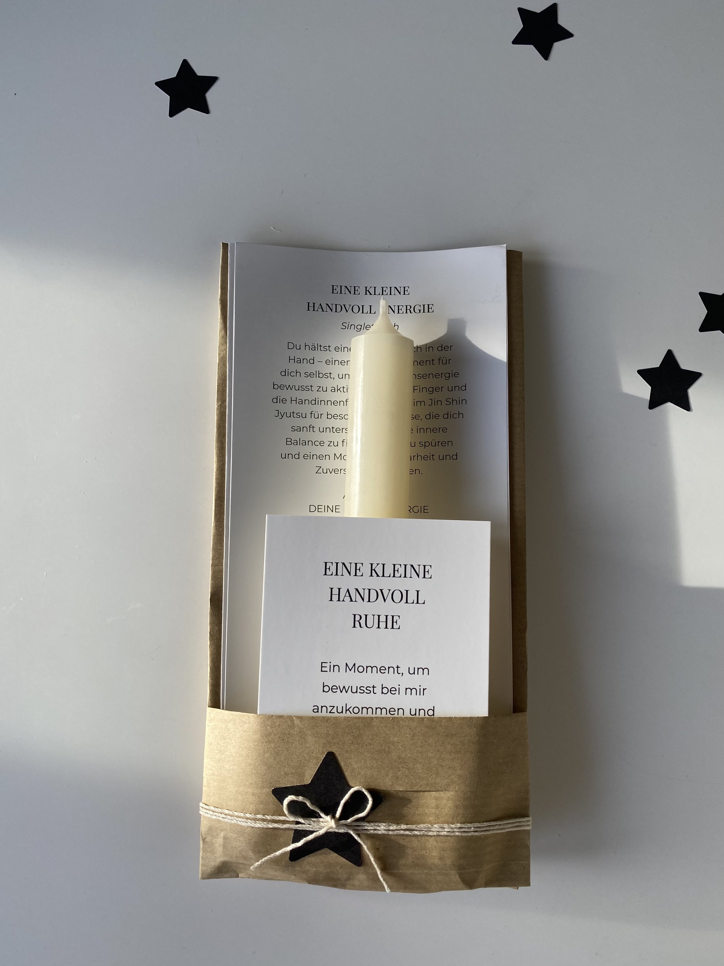Ein brauner Papierumschlag mit einer weißen Schnur, eine schwarze Sterne-Deko und ein weißes Papier mit dem Text 'EINE KLEINE HANDVOLL RUHE' und weiteren deutschen Texten. Darin befindet sich eine weiße Kerze, die auf einem weißen Blatt liegt, mit einem weiteren weißen Papier mit Text davor.