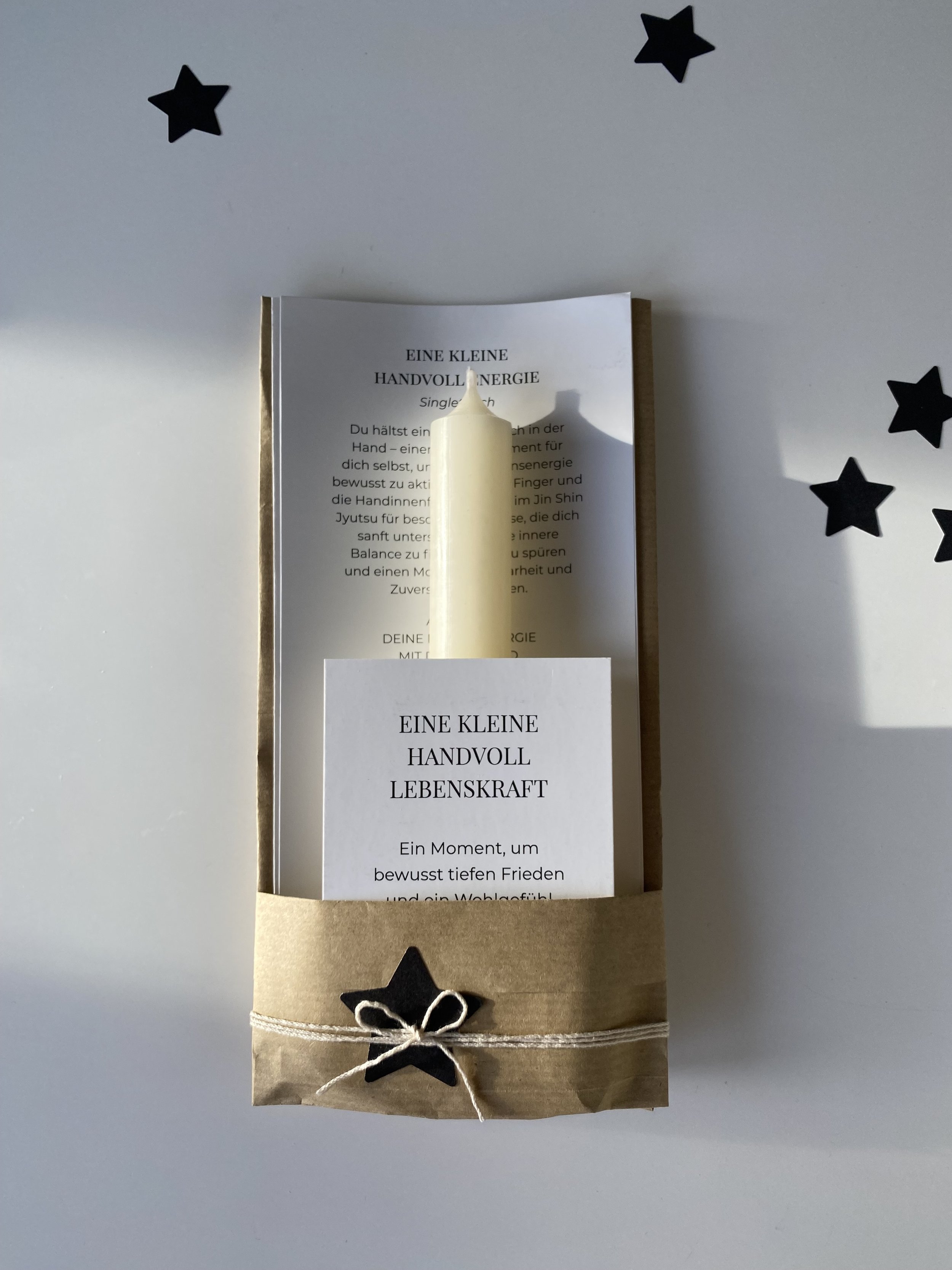 Beige Geschenkverpackung mit schwarzem Stern und weißen Band, enthält ein weißen Kerzenstift und gedruckte Texte in deutscher Sprache.