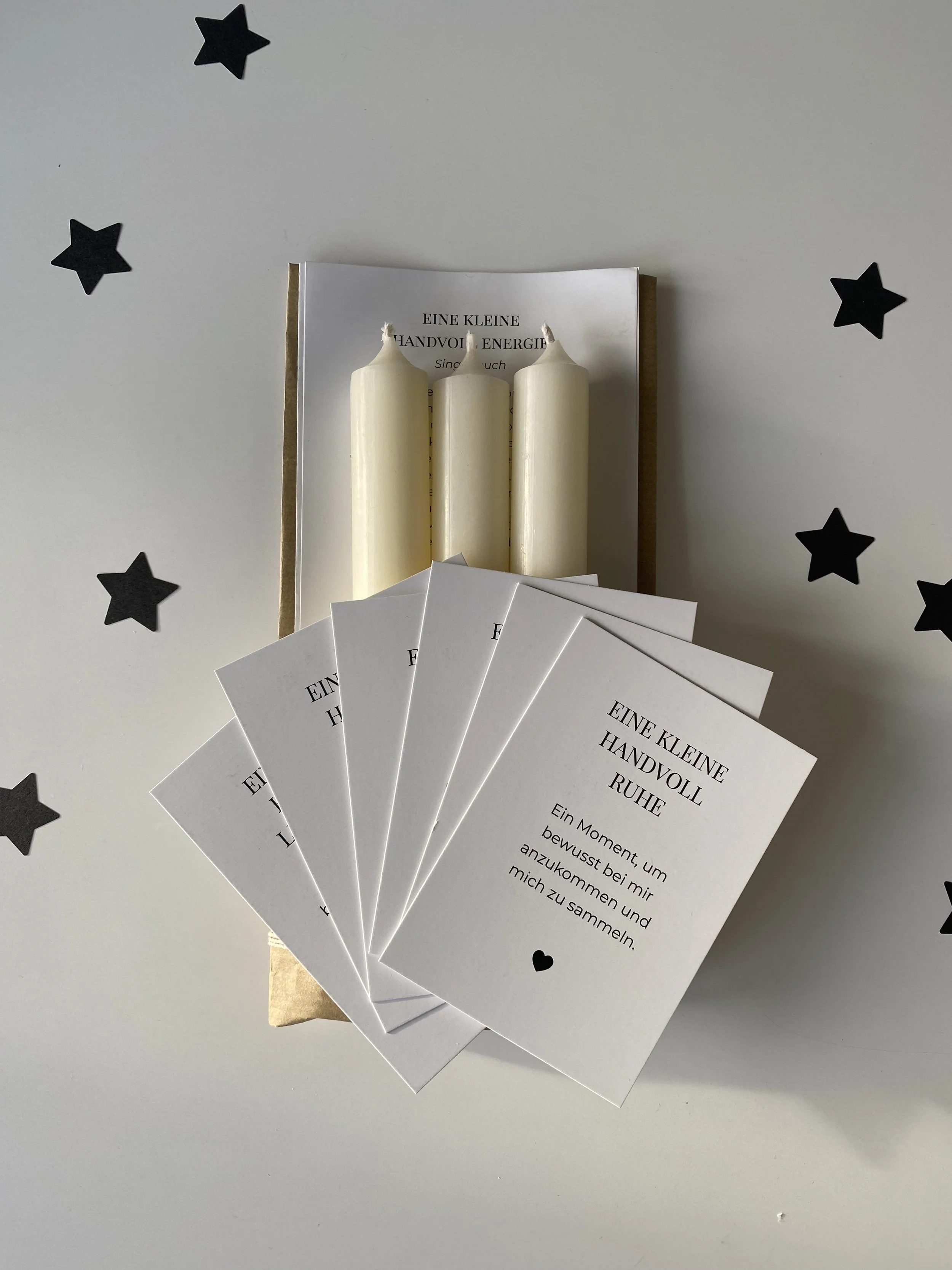 Eine kleine Handvoll weiße Kerzen in einer Geschenkbox, begleitet von mehreren weißen Karten mit der Aufschrift "EINE KLEINE HANDVOLL RUHE", auf einem weißen Tisch mit schwarzen Sternaufklebern.