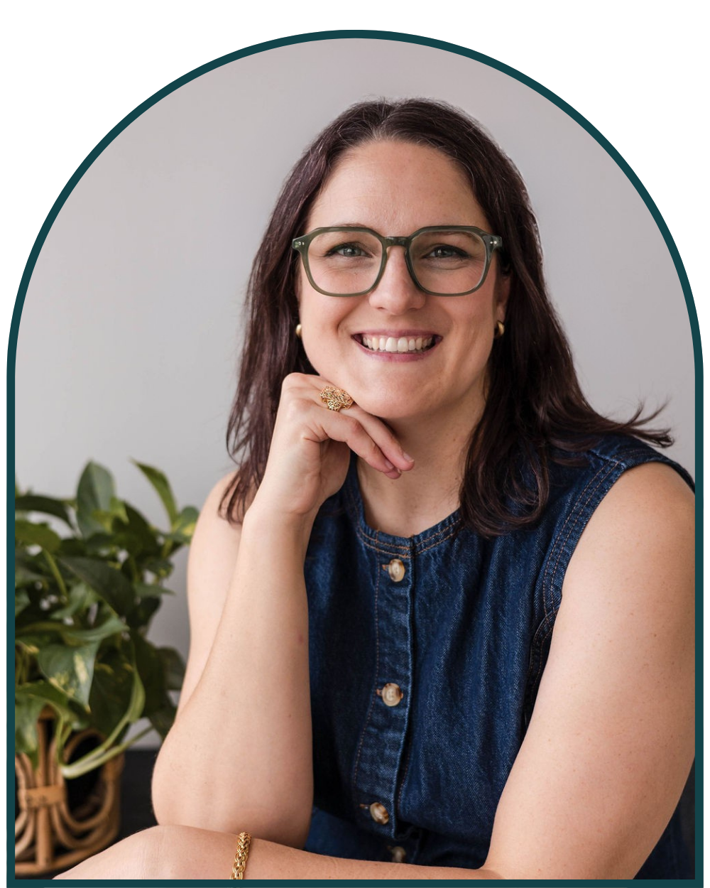 IFS therapist, Erin, sits smiling, leaning on elbow  wearing glasses and a blue denim shirt, with a green plant in the background.