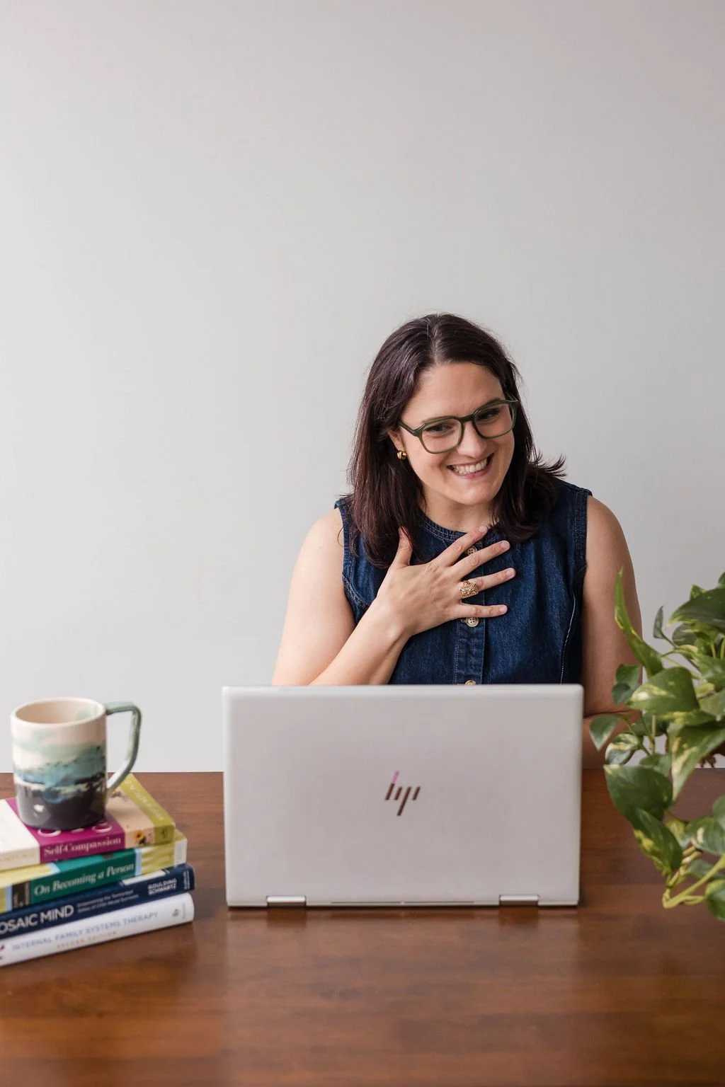 Erin Kehrier, LCSW, IFS and EMDR intensive therapist, smiling warmly at her desk with a plant, mug, and books nearby, hand on her heart, offering virtual intensive therapy in Texas and Michigan