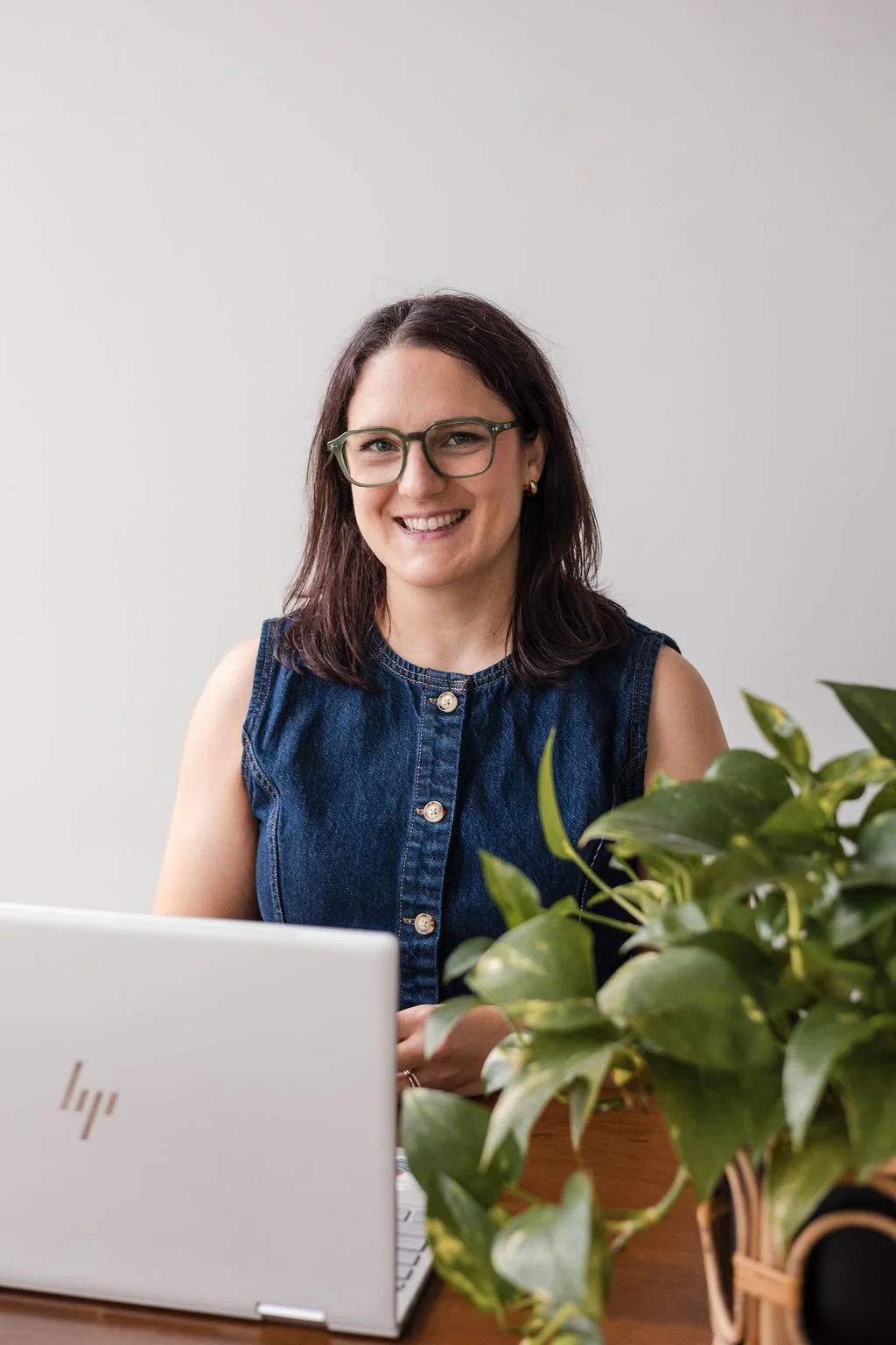 Erin a Adult ADHD Therapist with brown shoulder length hair and glasses and a denim vest sitting behind a plant and laptop smiles to the camera