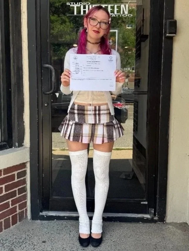 Young woman with pink hair and glasses standing outside a storefront, holding a certificate, wearing a plaid skirt, thigh-high white socks, and black shoes.