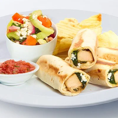 A photo of the spanikopita roll-up with strips of grilled chicken, blanched spinach, and crumbled feta wraped in a tortilla and grilled golden-brown on a plate with a side of potato chips, cucumber-tomato salad, and small bowl of tomato salsa
