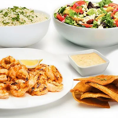 Grilled Shrimp Family Feast with a plate of grilled shrimp with lemon wedge, side of greek salad with lettuce, tomatoes, cucumber, and feta cheese, bowl of basmati rice with, and crispy baked pita chips