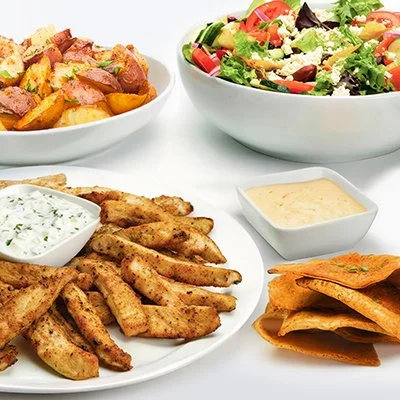 A photo of the Grilled Chicken Family Feast showing a plate of grilled chicken with taziki sauce, a dish of roasted potatoes, and a bowl of greek salad