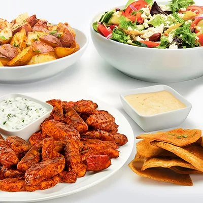 A photo of the spicy harissa chicken family feast, showing strips of grilled chicken covered in harissa sauce, a side of roasted potatoes, and a bowl of greek salad
