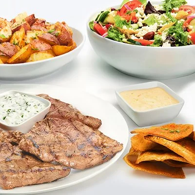 Chargrilled Lamb Family Feast with roasted potatoes, a Greek salad with tomatoes and cheese, and baked pita chips
