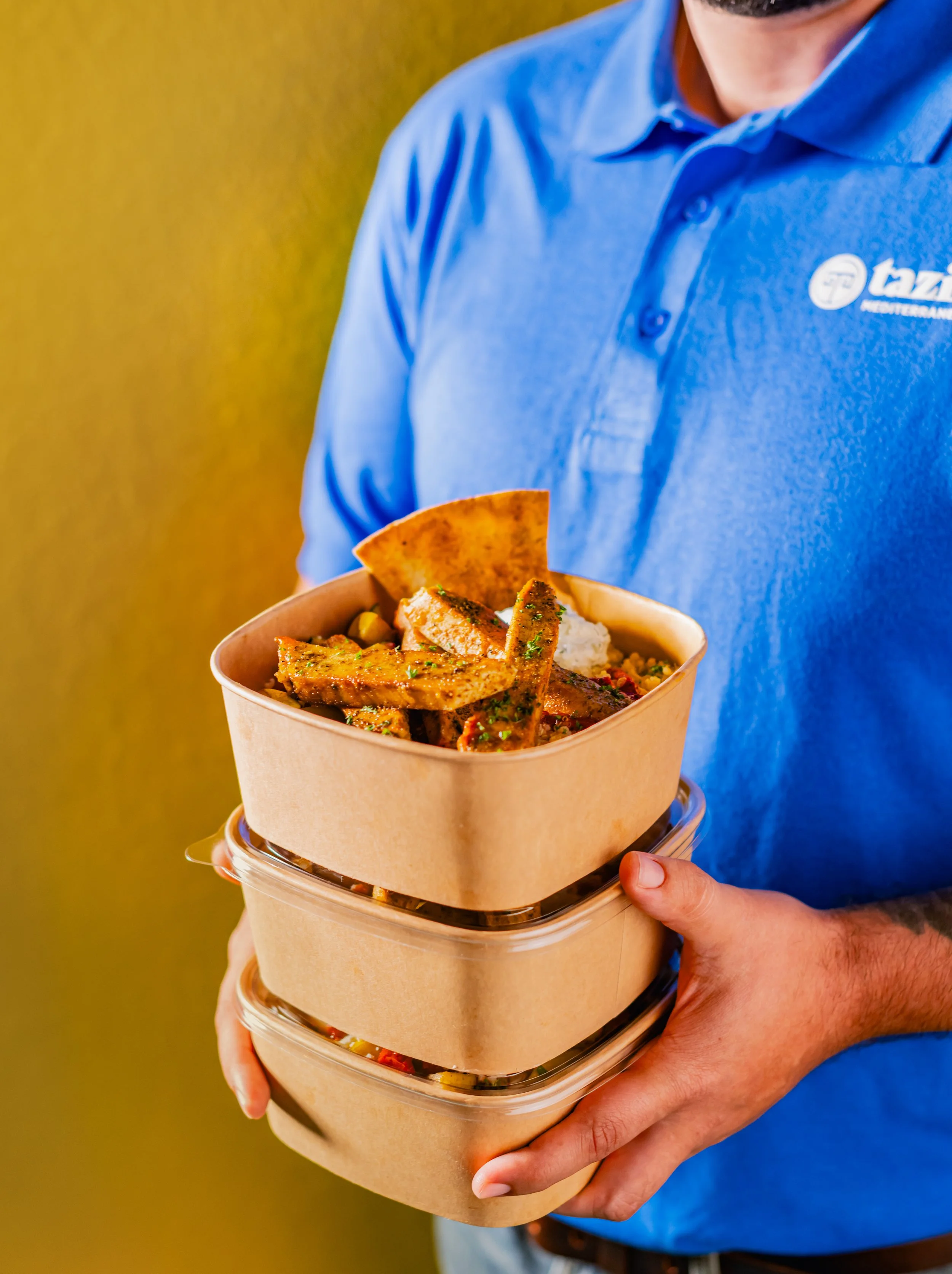 a photo of a man wearing a blue Taziki's polo, holding a stack of three brown paper bowls filled with delicious meals