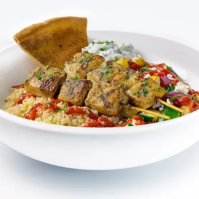 a photo of a white bowl filled with a bed of quinoa and topped with grilled veggies, crumbled feta, a drizzle of bright red harissa sauce and two chicken kebobs