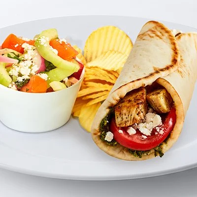 a photo of the grilled chicken basil pesto pita with strips of grilled chicken, sliced tomato, feta and basil pesto wrapped in a pita with a side of potato chips and cucumber salad