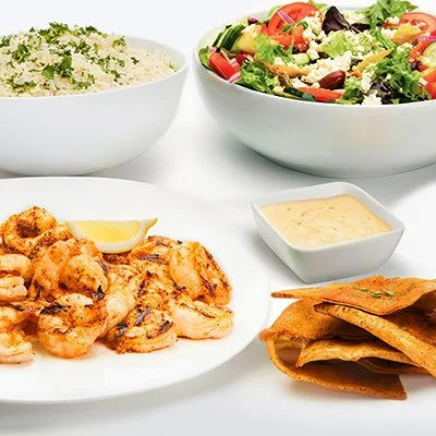 Grilled Shrimp Family Feast with a plate of grilled shrimp with lemon wedge, side of greek salad with lettuce, tomatoes, cucumber, and feta cheese, bowl of basmati rice with, and crispy baked pita chips