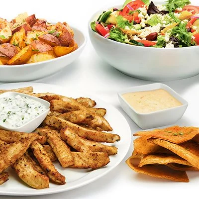 A photo of the Grilled Chicken Family Feast showing a plate of grilled chicken with taziki sauce, a dish of roasted potatoes, and a bowl of greek salad