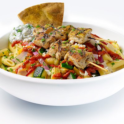a photo of a white bowl filled with penne pasta and topped with grilled veggies, crumbled feta, shaved parmesan, a drizzle of bright red harissa sauce and two grilled chicken kebobs