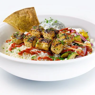Bowl of grilled chicken kebabs with vegetables, rice, flatbread, and sauce, garnished with herbs.