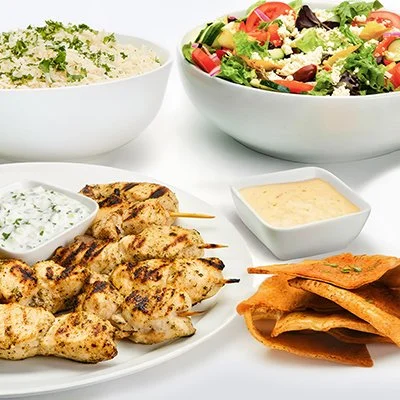 Chicken Kebob Family Feast, with a plate of chicken kebobs and taziki sauce, a bowl of fresh salad, a bowl of rice, and baked pita chips.