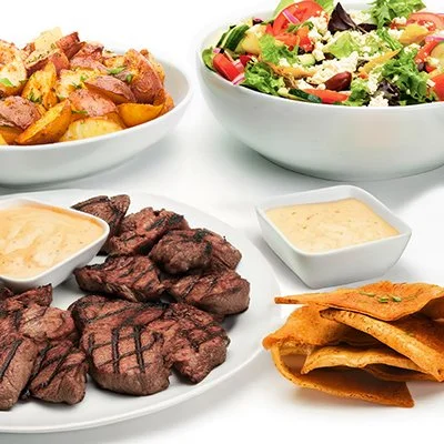 a photo of the Grilled Beef Family Feast showing a plate of grilled beef medallions with horseradish sauce, a bowl of roasted red potatoes, and a bowl of greek salad