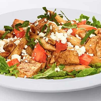 A photo of a white plate filled with a bed of mixed lettuces with penne pasta and grilled chicken, tossed in balsamic vinaigrette, topped with tomatoes, feta, and fresh basil. Served with a baked pita chip.