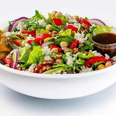 a white bowl is filled with Mediterranean salad, showcasing chickpeas, red onion, roasted red peppers, candied pecans, and crumbled feta with balsamic vinaigrette