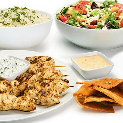 Chicken Kebob Family Feast, with a plate of chicken kebobs and taziki sauce, a bowl of fresh salad, a bowl of rice, and baked pita chips.