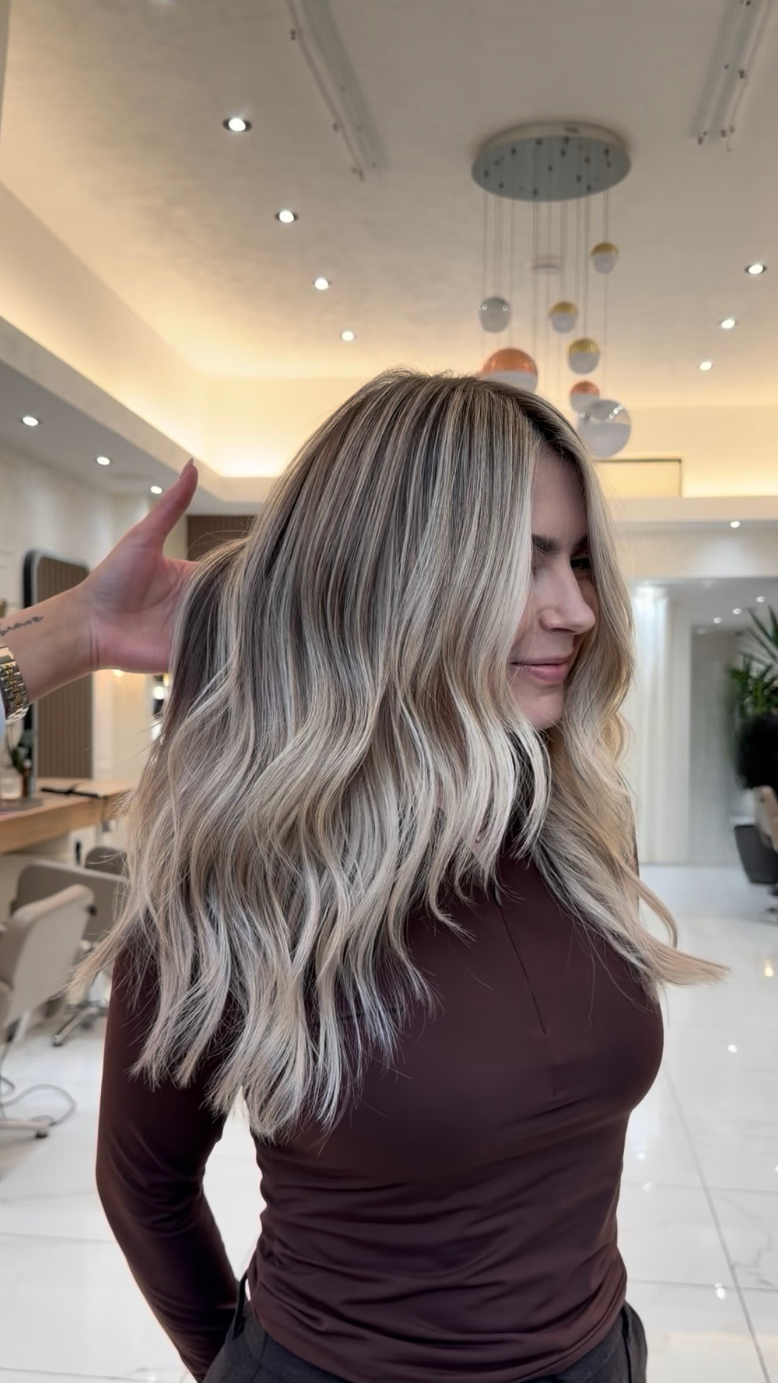 A woman with long, wavy blonde hair with darker roots, wearing a dark maroon long sleeve top, standing in a modern salon with a decorative ceiling light fixture.