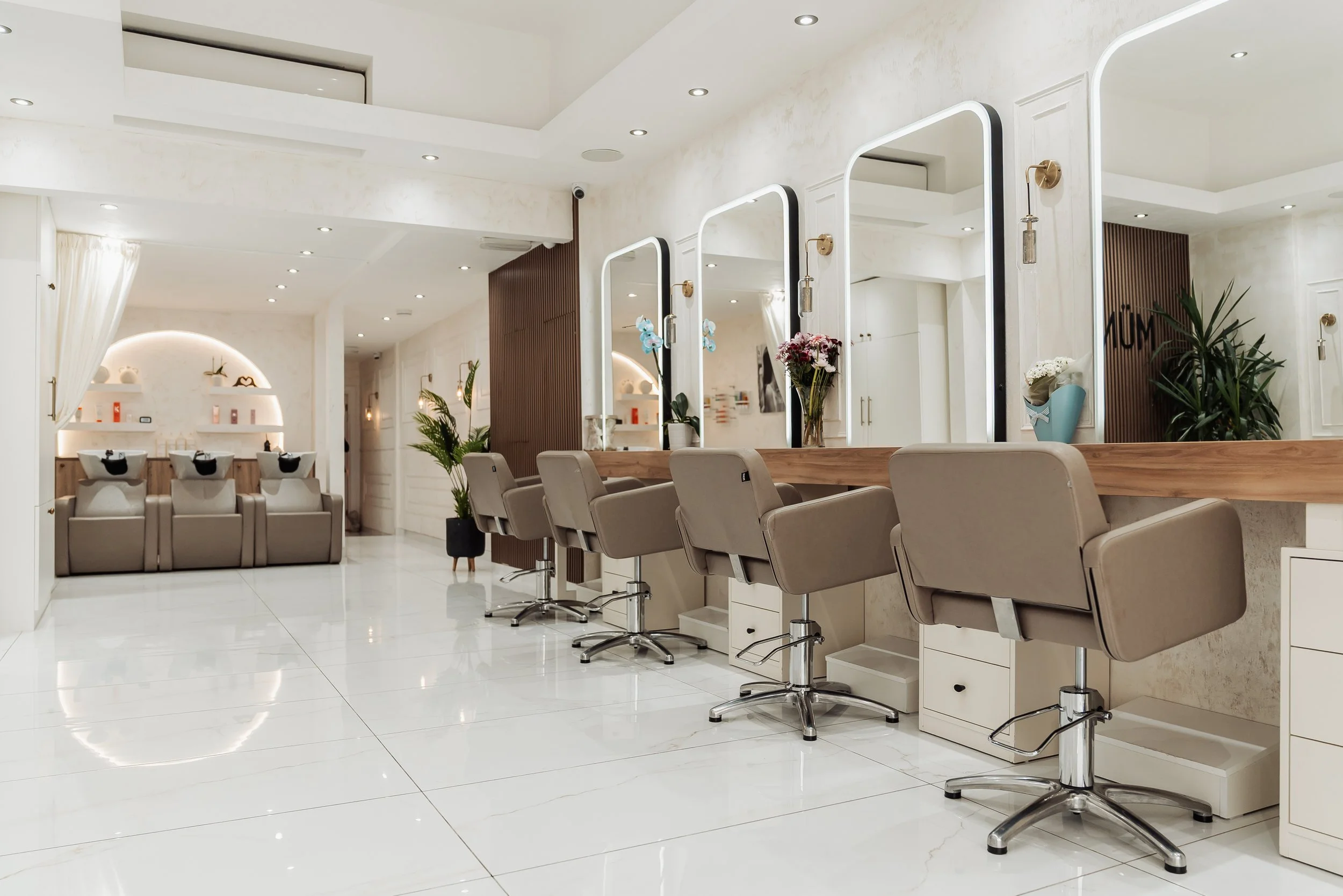 Modern hair salon with beige chairs, large mirrors, flowers, plants, and white walls with recessed lighting.