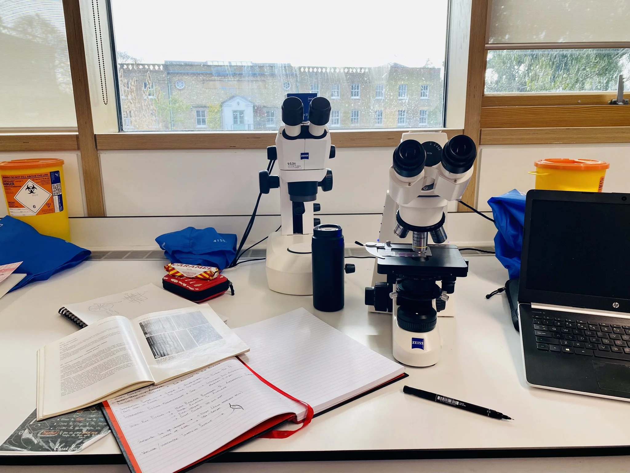 Scientific laboratory workspace with two microscopes, a laptop, and open notebooks on a white table near a window.