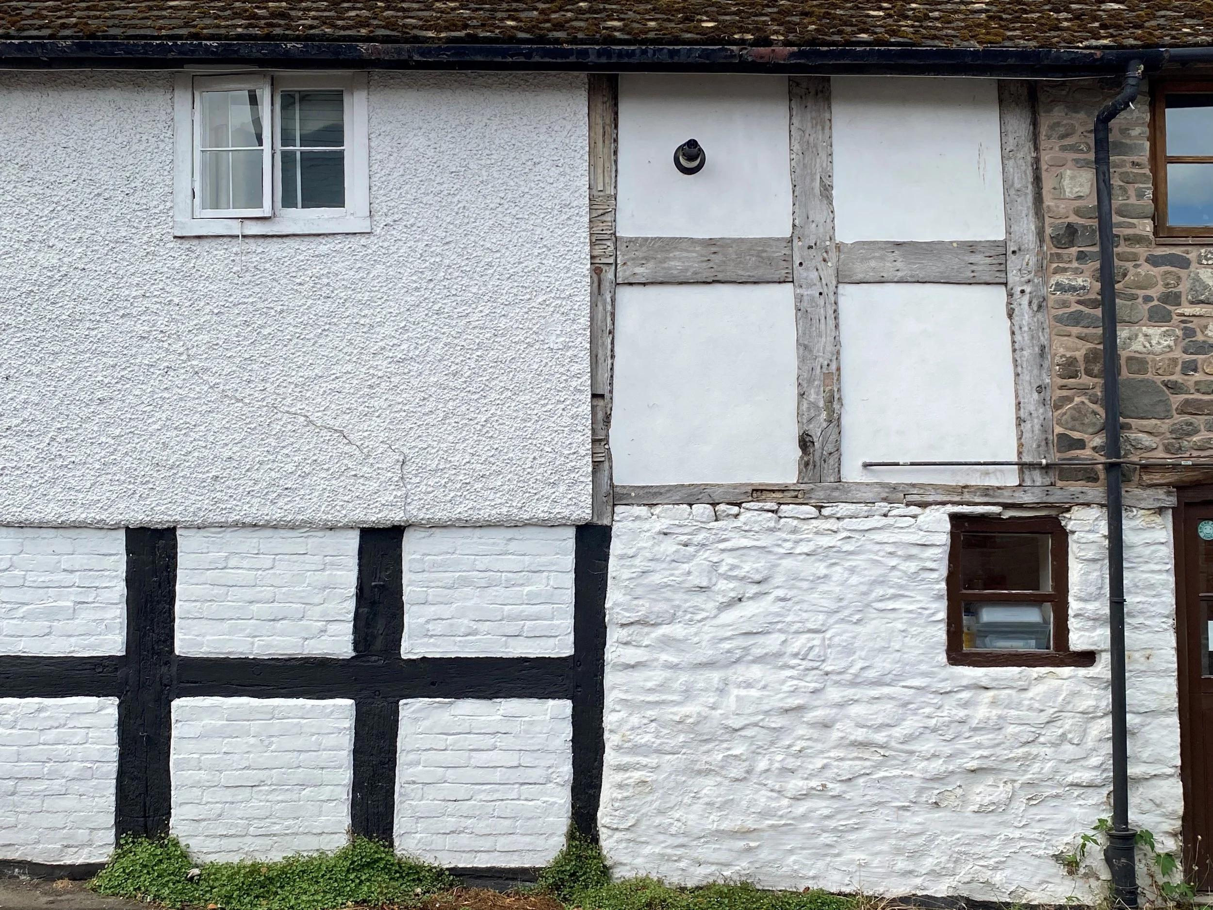 An historic half-timbered house with white walls, black wooden beams and stone accents on the right side, featuring a small window with a wooden frame and a drainpipe on the right edge, set against a background of an overcast sky.