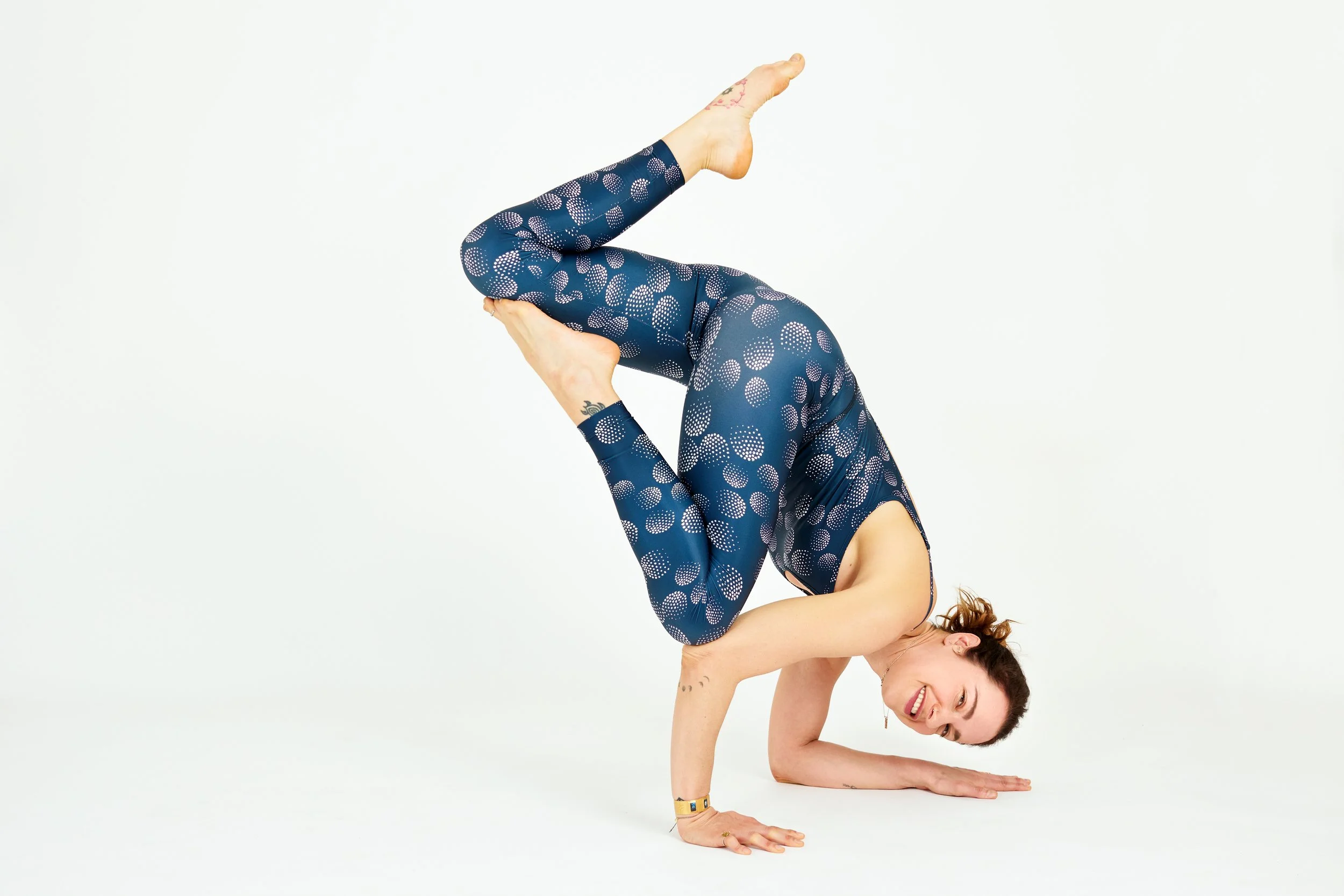 Insegnante di yoga in equilibrio su avambracci e mani, sorride mentre esegue una posizione avanzata in abbigliamento tecnico blu con motivi a cerchi, su sfondo bianco.