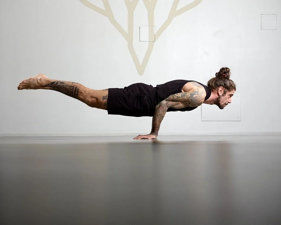 Insegnante di yoga esegue la posa avanzata Mayurasana (posizione del pavone) su un pavimento chiaro, indossando un completo nero e mostrando tatuaggi su braccia e gambe.